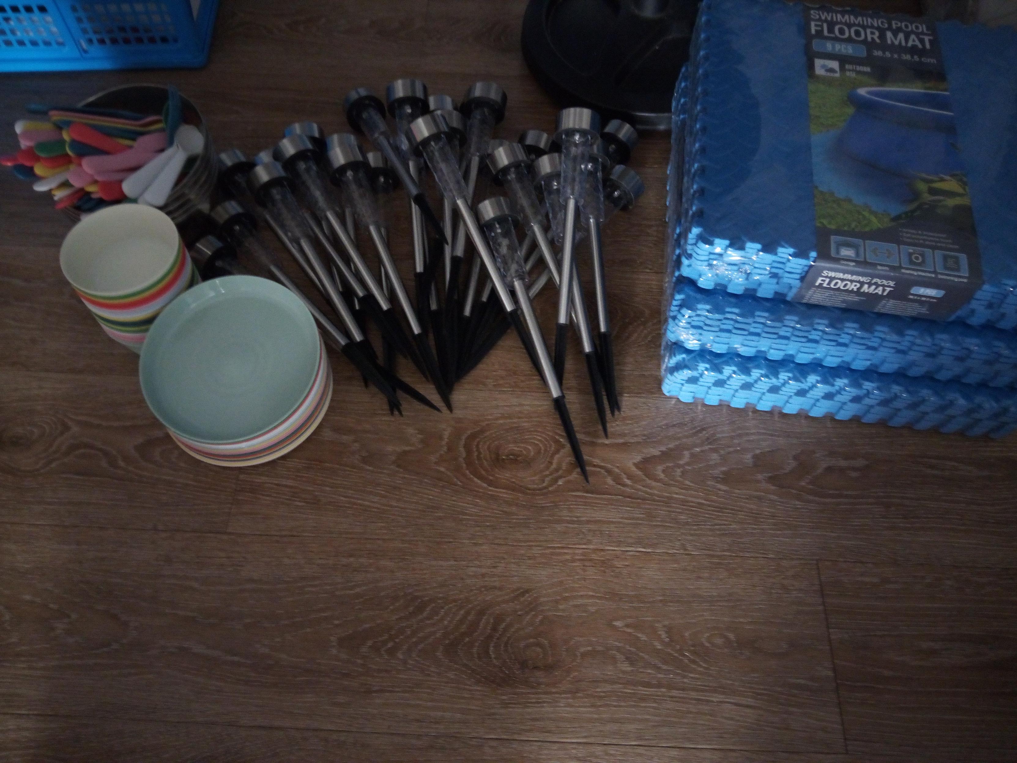 Various items on the floor; 3 packs of rubber floor mat tiles, a large pile of solar-powered lights that you stick into the ground, a stack of reusable plastic plates, a stack of reusable plastic bowls, and a few stainless steels bowls containing a mountain of reusable plastic cutlery